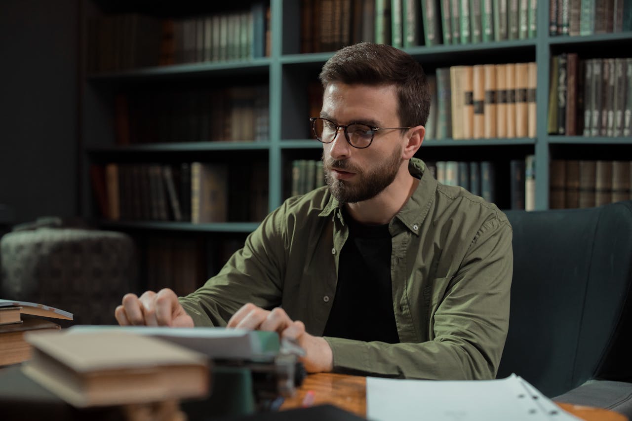 A young man with glasses typing on a vintage typewriter in a library.