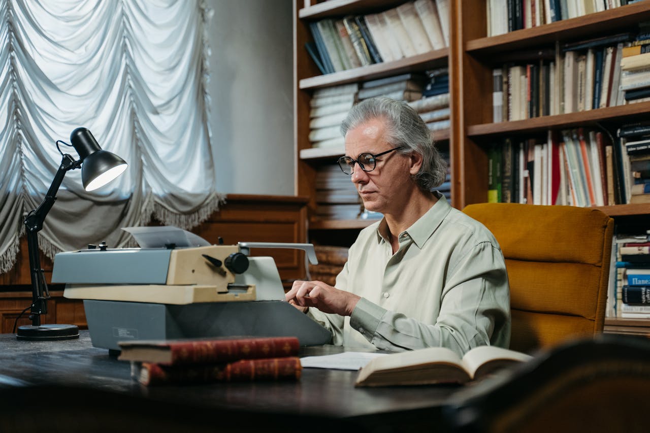 Senior man with glasses and gray hair typing on vintage typewriter in cozy library setting.